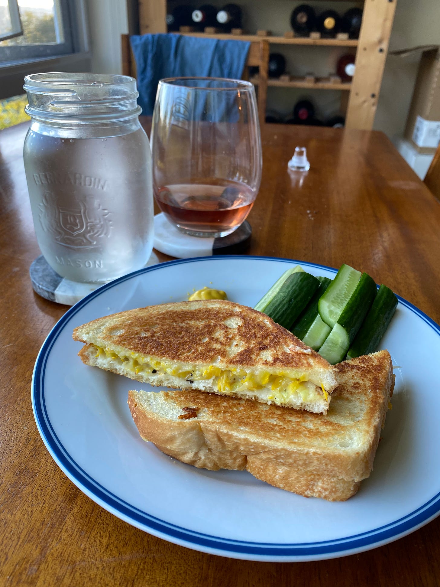 A grilled cheese sandwich with shredded yellow zucchini, sliced in half and stacked on a plate next to some cucumber pieces and a little bit of mustard. In the background, a glass of rosé wine is next to a mason jar of cold water.