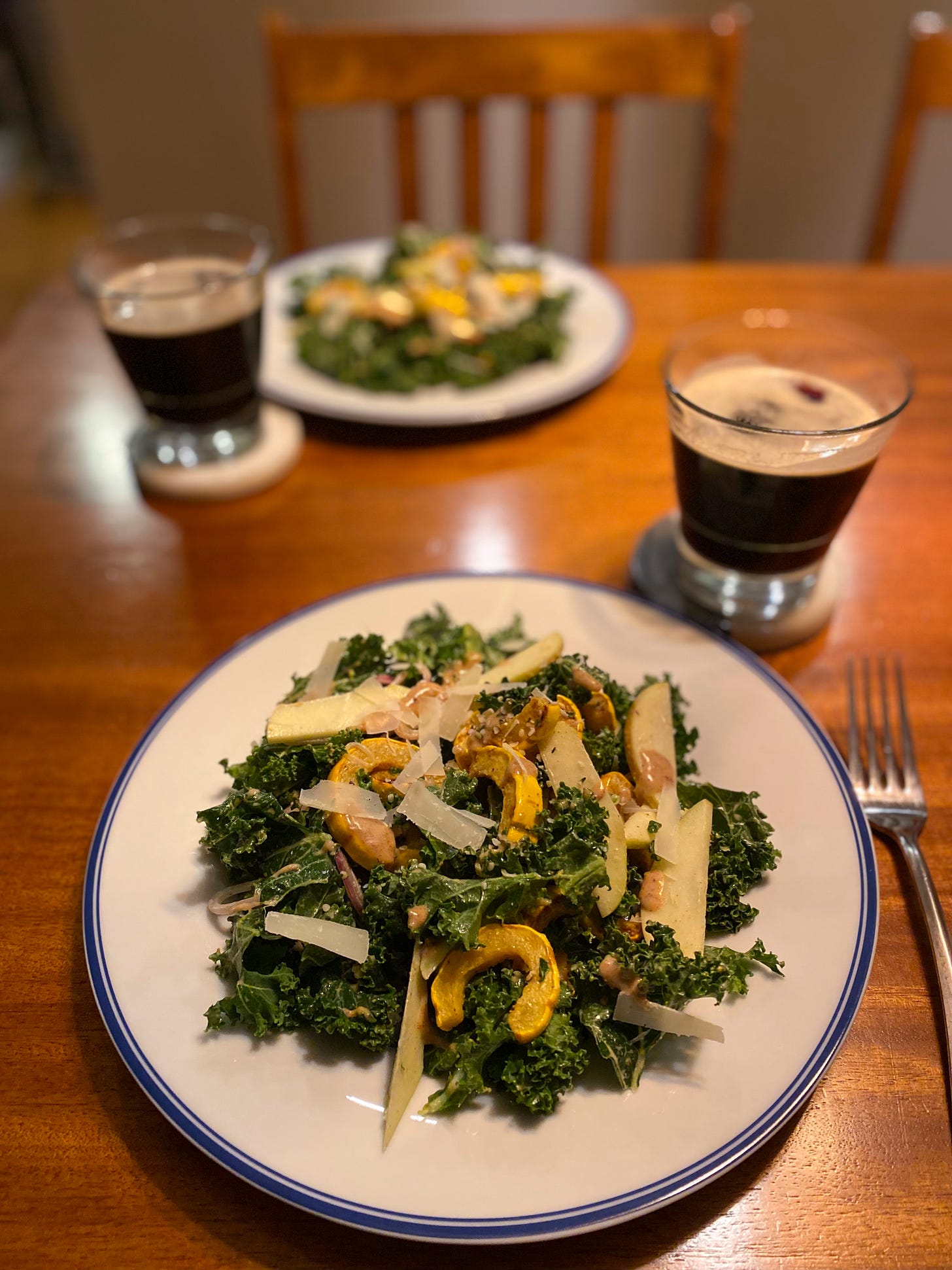 Two plates of kale salad across from each other on the table, each with a glass of dark beer on a coaster beside it. The salad has slices of apple and roasted delicata squash, and pieces of pecorino and hemp seeds scattered overtop.