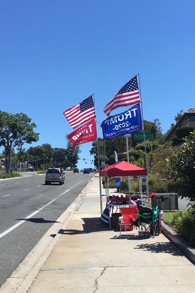 photo of a pop-up booth on a sidewalk with two towering American flags and two large Trump 2020 flags 