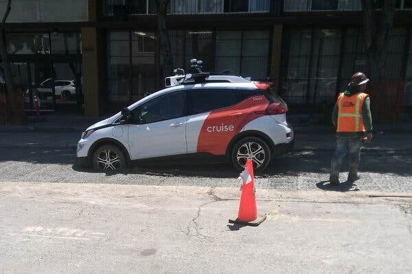 A white and red, driverless Cruise car with its front wheels stuck in wet concrete. A person in an orange safety vest stands next to the vehicle, near a traffic cone.