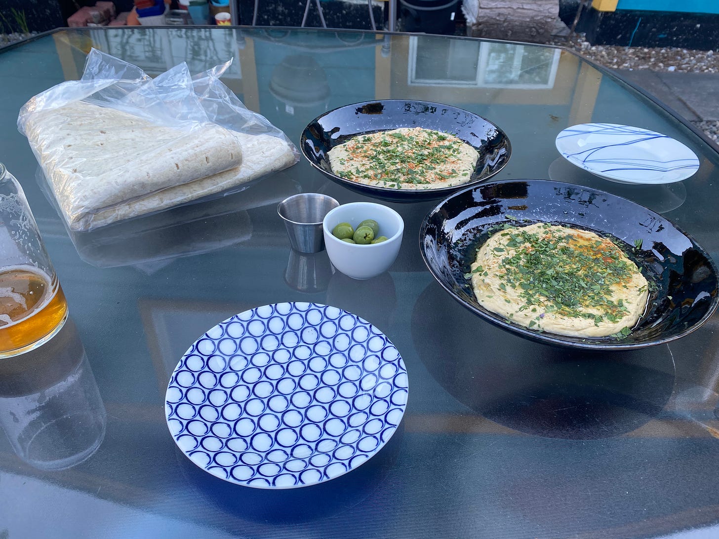 On an outdoor glass table, two shallow black bowls with hummus and baba ganouj, dusted with smoked paprika and lots of herbs. They are flanked by two small side plates, and next to them is a small dish of green olives and a package of taftoon.