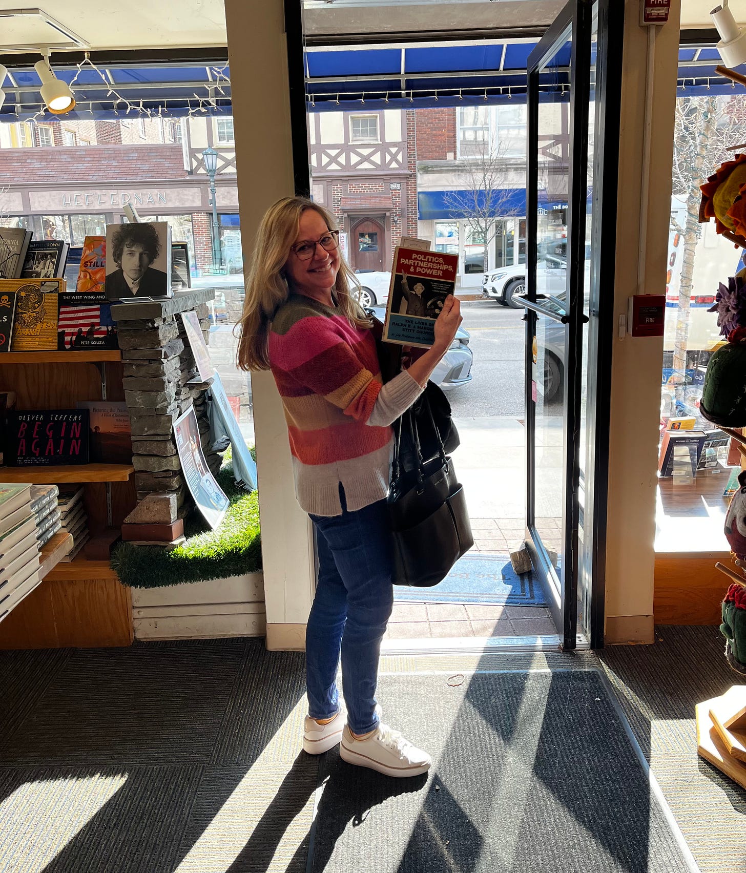Memoir Coach Christine Wolf stands in the doorway of a bookstore holding her debut novel, Politics, Partnerships, & Power: The Lives of Ralph E. and Marguerite Stitt Church. Wolf is a blonde woman wearing glasses, a striped sweater, and denim jeans. 