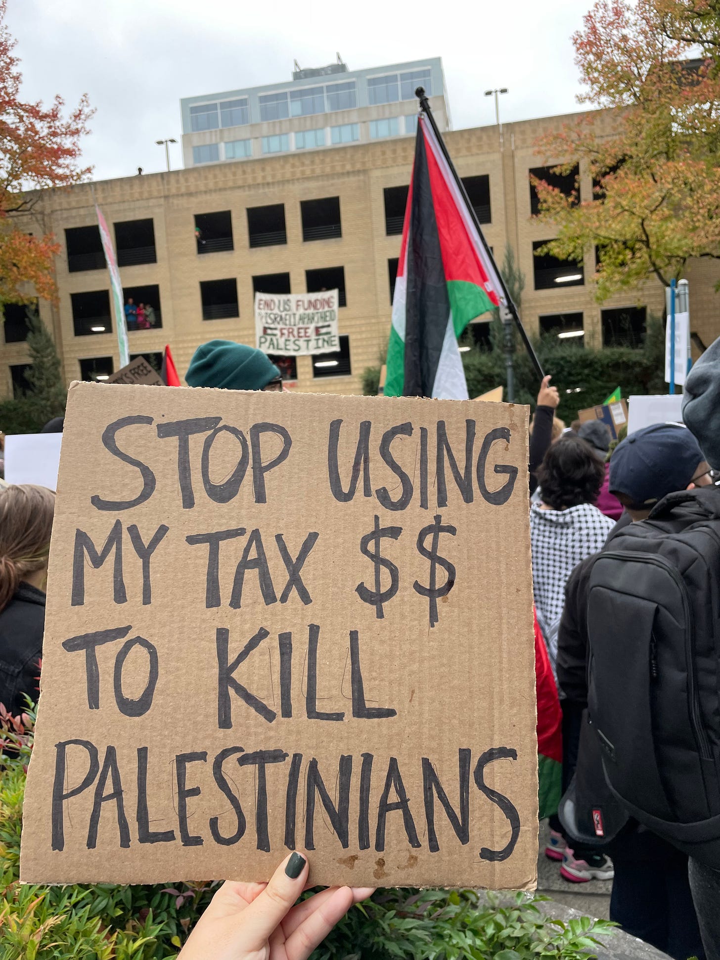 outside in POrtland ORegon in front a huge parking structure, people are wearing hats and beanies and holding up signs and palestinian flags. A cardboard sign is in the foreground and it says stop using my tax $$ to kill palestinians