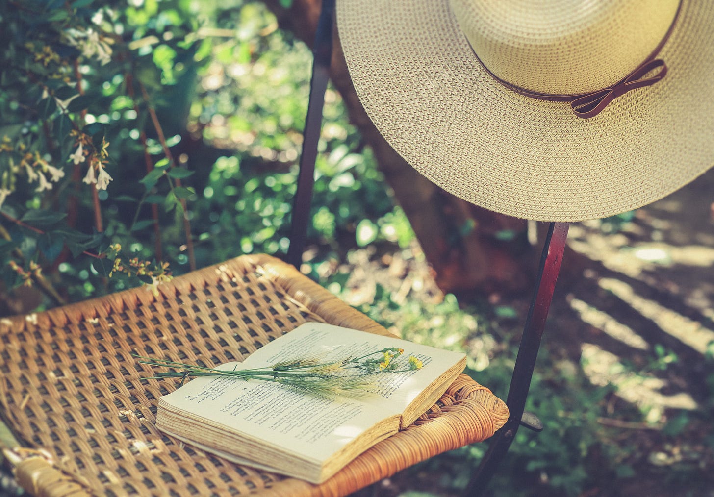 Wicker chair in a garden. There’s an open vintage book over the seat, and a few sprigs of yellow flowers over it. A summer hat hangs from the chair’s back.