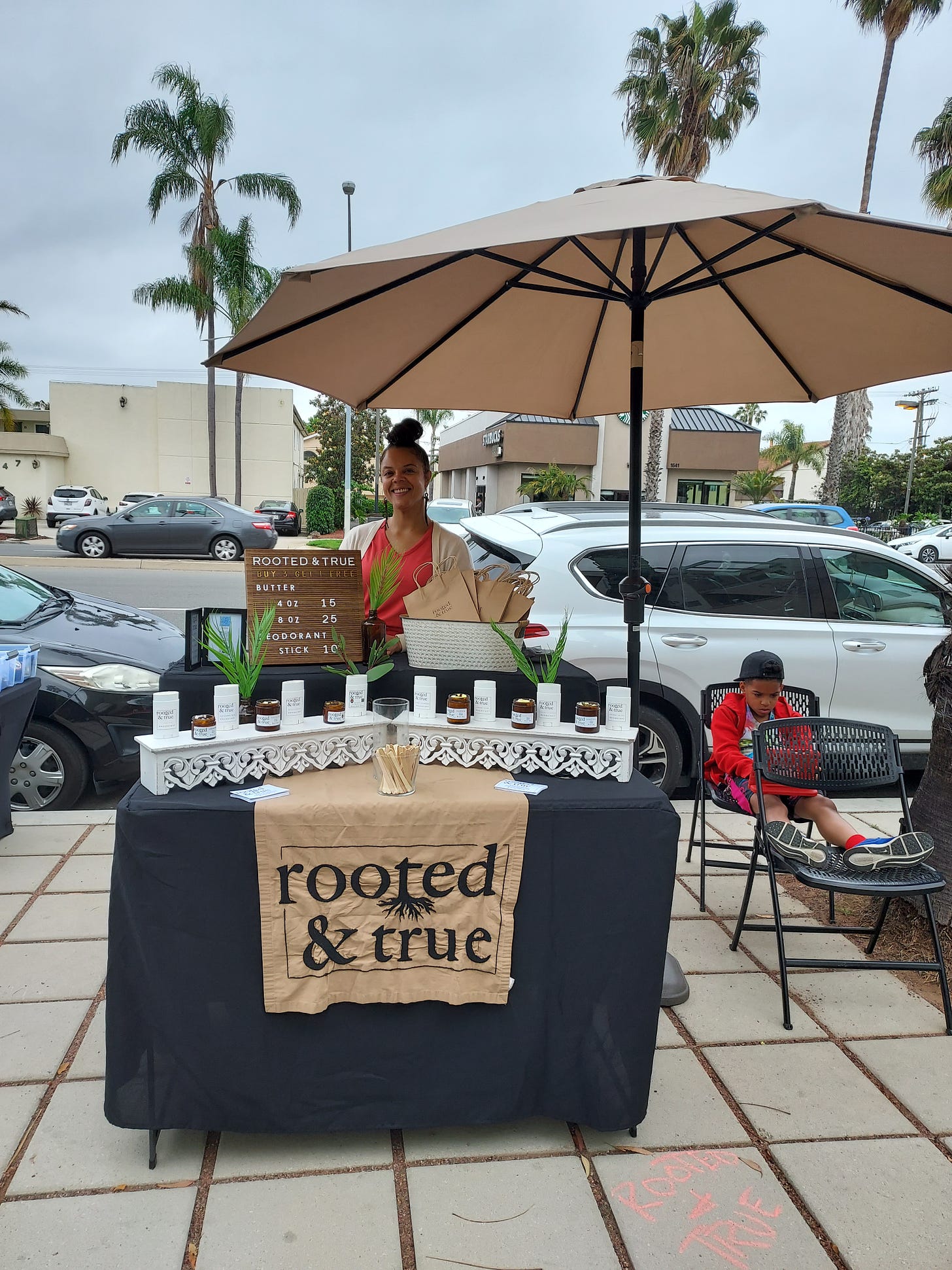 A woman poses behind a marketplace table set up with deoderant and body butter under a beige umbrella on the sidewalk. Palm trees dot the background.