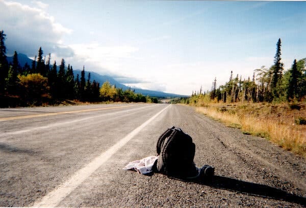 A black backpack rests on the side of an empty road lined with trees and yellowing grass.