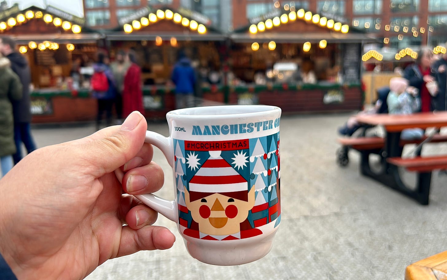 A hand holds a cup that says ‘Manchester’ and ‘#MCRChristmas’, with brightly lit huts selling Christmas drinks in the background.