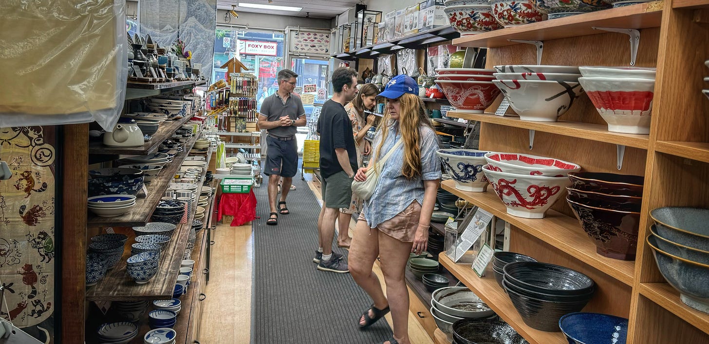 The housewares aisle inside Sanko. On the shelves are a variety of patterned bowls and dishes. All of the shoppers are White.