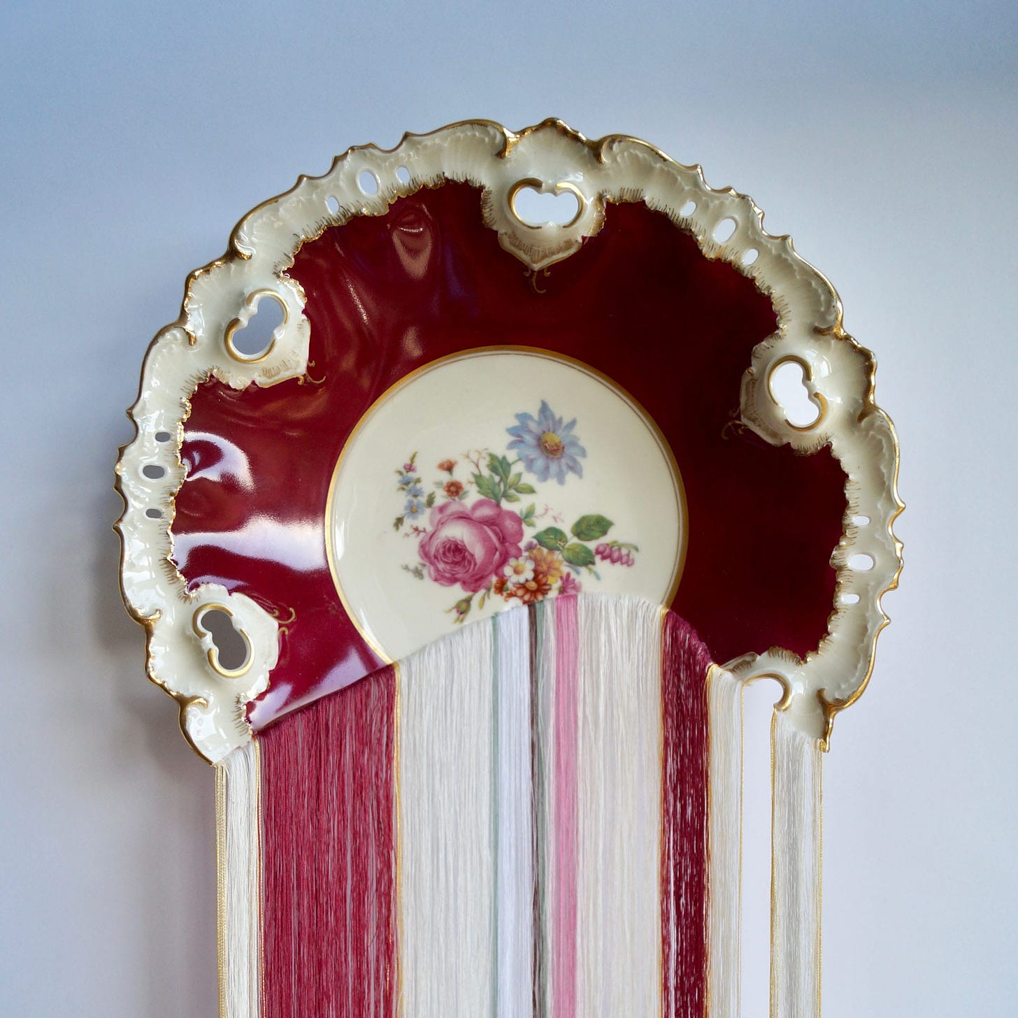 Hundreds of filigree threads hang from a broken porcelain plate in hues of reds, whites, and pinks.