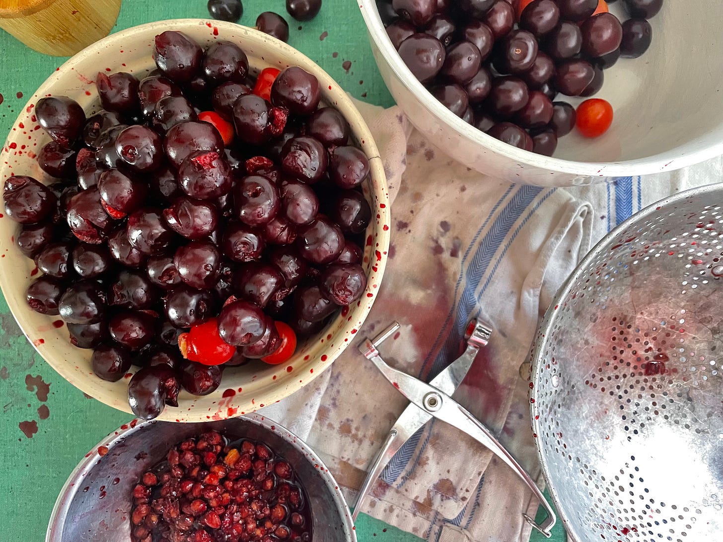 A scene of pitting cherries. There is a yellow bowl near the center with pitted cherries. There’s a white bowl partly out of frame with whole cherries. There’s also a bowl of cherry pits, a colander, and a handheld cherry pitter. All of these atop a Kelly green counter that has a cotton towel on it. Cherry splatters and stains everywhere.
