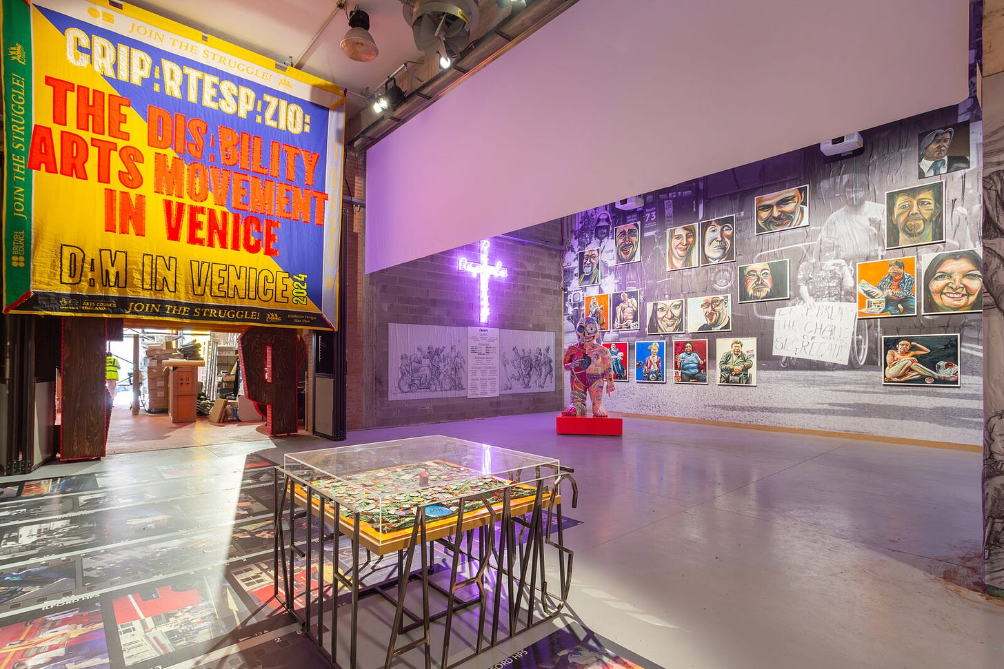 A spacious and tall gallery space has a lot going on. A huge banner with the title of the exhibition in yellow and red and blue. A squiggly Christian cross in glowing neon purple. Portraits on a wall, images on the floor.