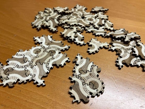 A heap of oddly-shaped puzzle pieces made of laser-cut plywood, with decorative patterns engraved on the top, some assembled to patches of a couple pieces. There are only two shapes, but their meandering edges fit together in various different ways that make the engraved patterns match up.