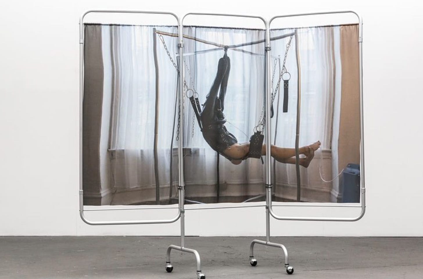 A photograph is printed on a wheeled 3-panel privacy screen that you might encounter in a clinical setting. In the image, a person is suspended in a leather sling with their hands tied over their head.