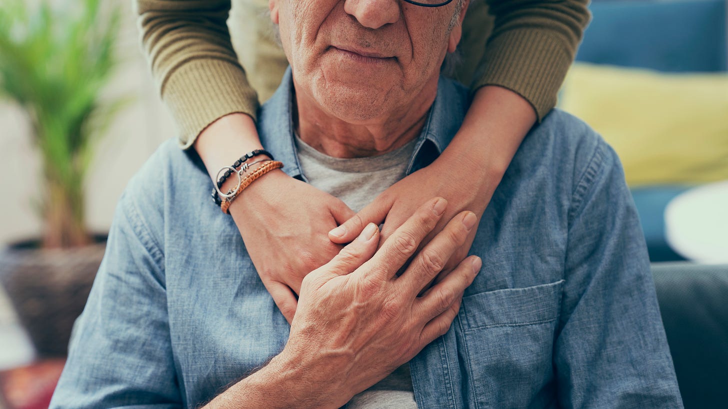 Older man, seated, visible from nose to chest. Arms of younger woman visible on his shoulders.