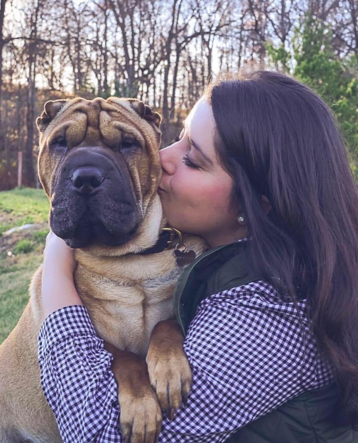 A caucasian woman in her late thirties with long brown hair is shown in profile view kissing her Shar Pei dog. The dog is looking directly at the camera with a big black nose and muzzle surrounded by a wrinkly face of tan fur