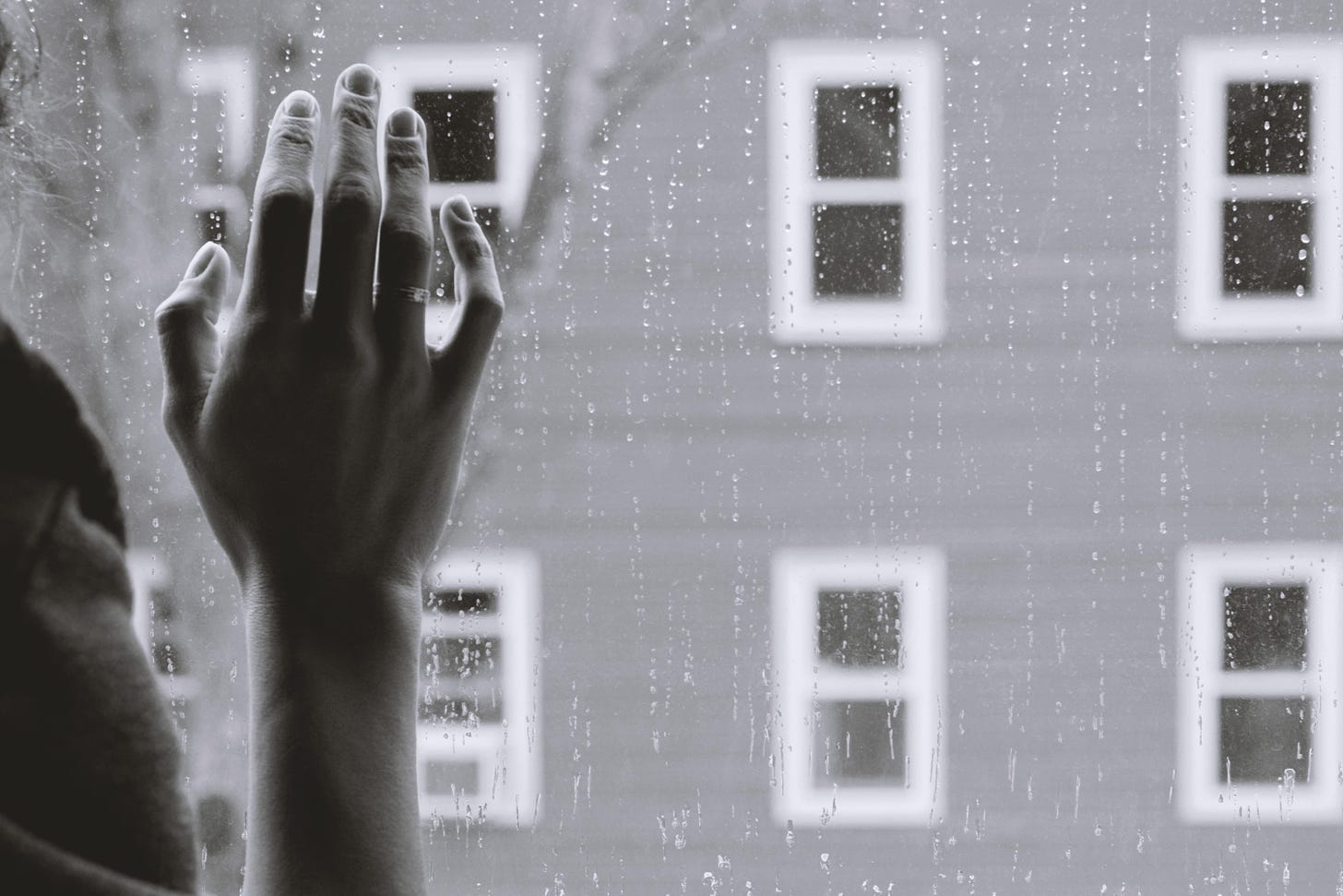 Grayscale photo of a hand raised against a window pane with droplets of rain and a building with rows of windows in the background.
