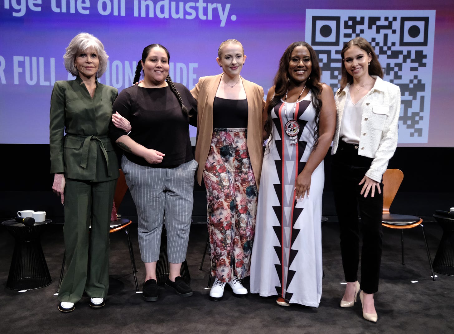 Jane Fonda stands next to four other women on a stage, where all five women pose for a photo. Behind them are wood chairs and a purple screen with a QR code.