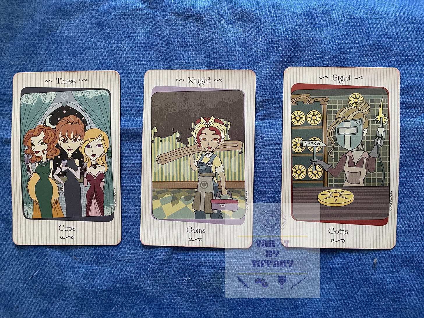 3-card reading using the Vanessa Tarot. Cards (L-R): 3 of Cups, Knight of Coins, 8 of Coins. The cards are laid out on a blue cloth background.