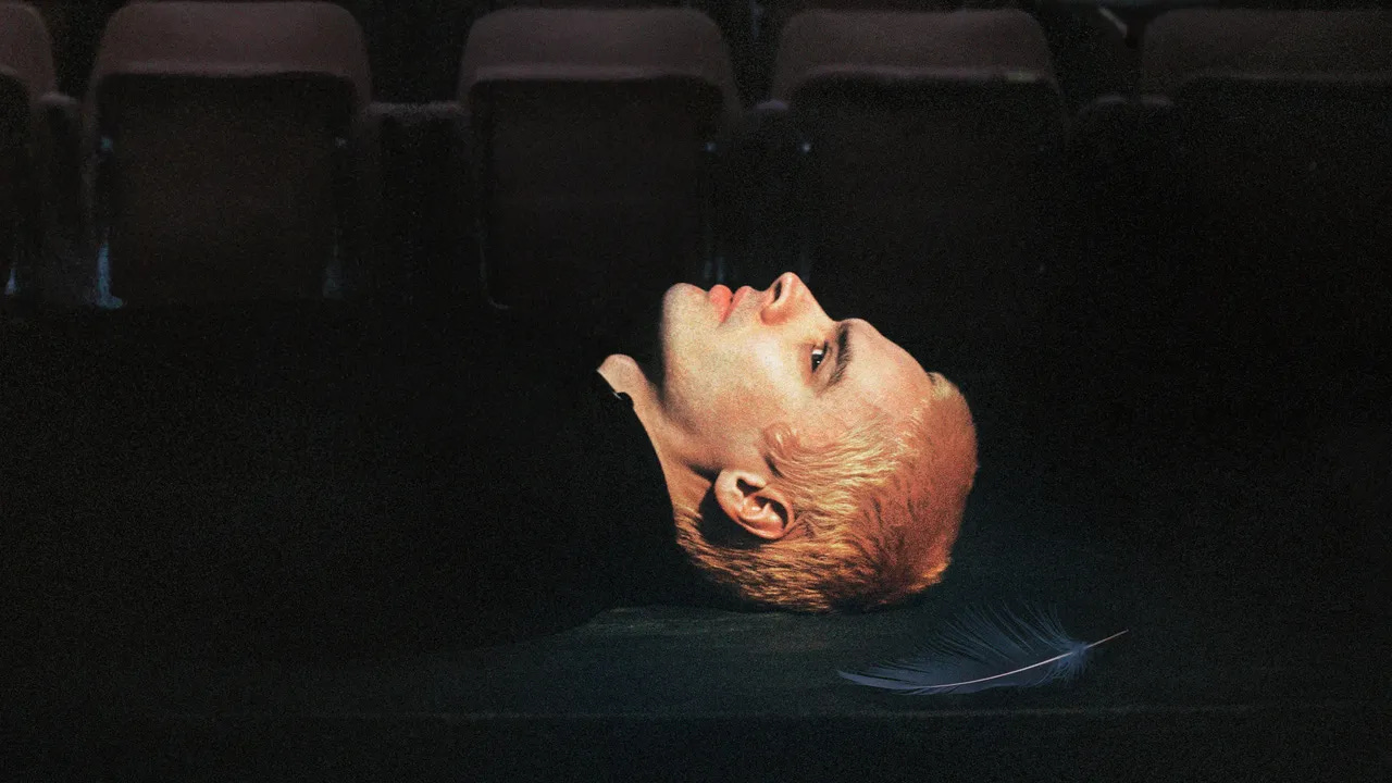 A person in black with short dyed blonde hair lies horizontally in a dark room. A row of cinema-style seats are visible in the background. A single feather lies by their head.