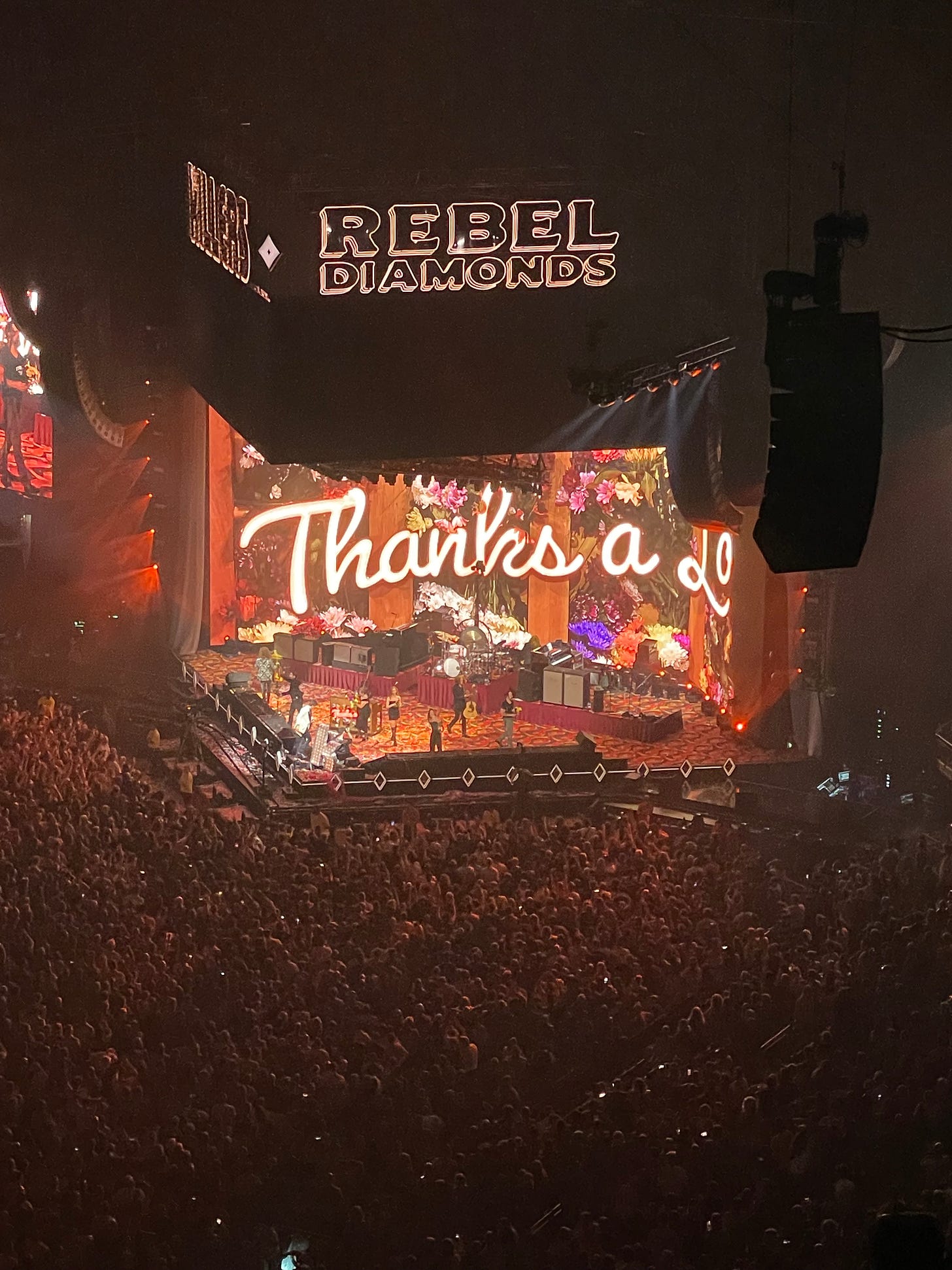 The Killers play on a triangular stage in the O2 arena in London. Their backdrop reads “Thanks a Lot”, and their name, alongside their latest album, “Rebel Diamonds” are lit up on signs above the stage.