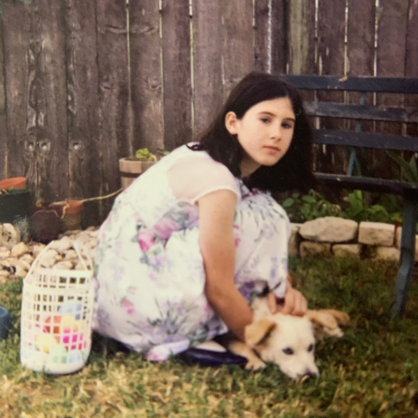 Young Lyric, with medium length black hair, blank faced, crouched, blank faced, in a pastel easter dress. They are petting a small tan puppy, that is laying in the grass. On the other side of them is a small white plastic laundry basket of multi-colored plastic easter eggs.