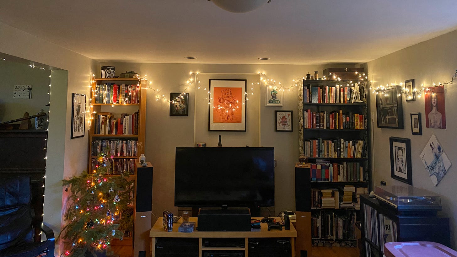 My living room, with small white lights hung loosely around the wall, over the framed pictures, and across the bookshelves, which flank the tv on its stand in the centre below the mantel. To the left of the tv is a small tree decorated with coloured lights, a little more than half the height of the bookshelf it's in front of. More lights are visible on the doorway to the dining room on the left, and the Radiohead album 'OK Computer' rests on pins above the record player on the right side of the room.