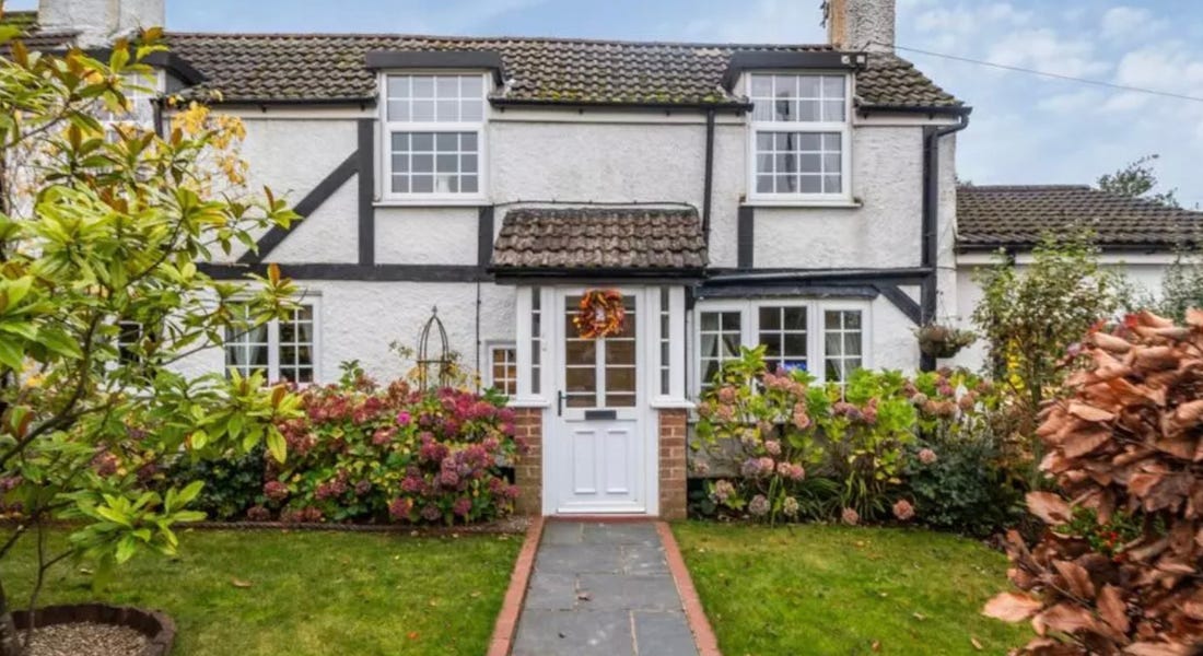 A white and black cottage with a door with a wreath, a grey path leading up to it, and lawns either side