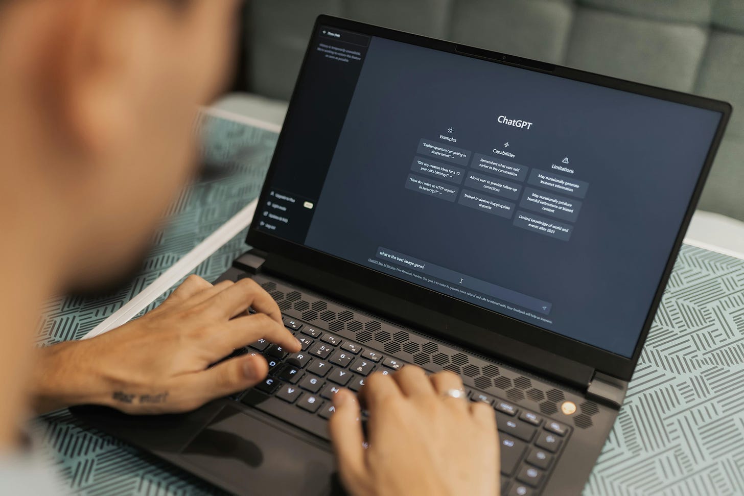 A visual of a laptop, screen showing the ChatGPT home screen, set of hands on the keyboard of the laptop. 