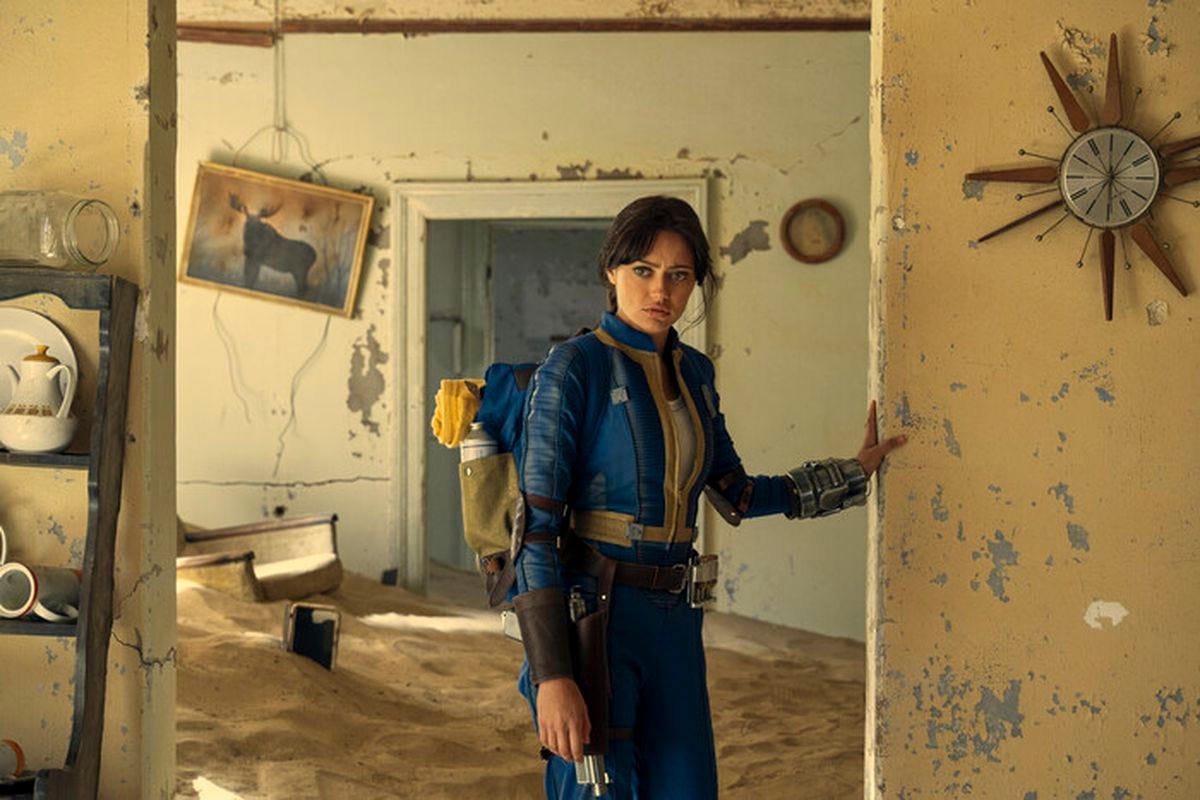 A young woman wearing a backpack stands in the doorway of a derelict room that appears to be abandoned. Wall decorations hang askew and the wall paint is chipped and damaged. The ground is covered in sand.