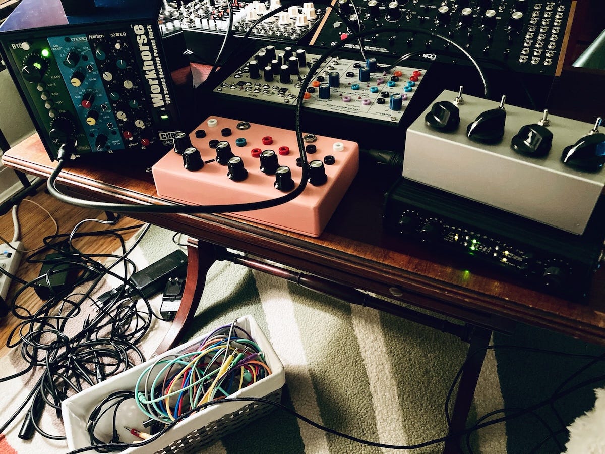 A small vintage coffee table crowded with small modular synthesizers and audio gear. A basket of cables and tangle of cords is under the table on a green and white striped rug.