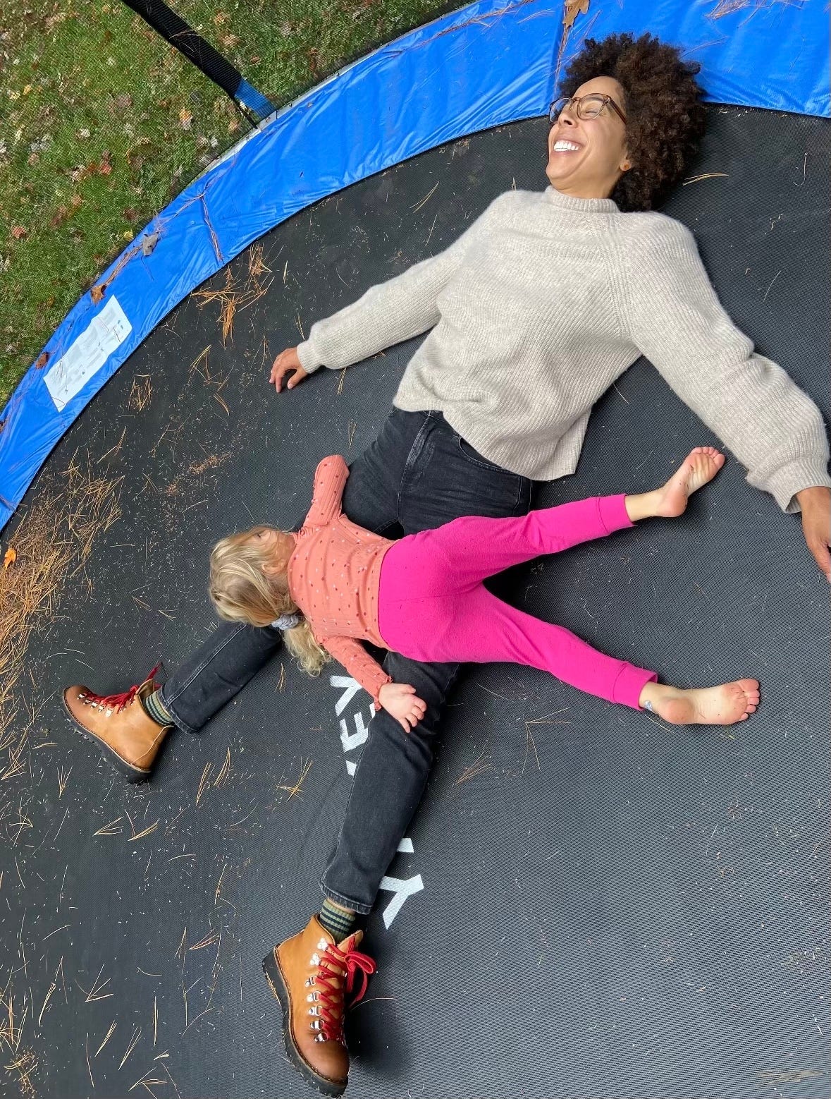 Ayana and Marlow lie on a trampoline outside. Marlow is lying across Ayana, her face is not visible. Ayana’s eyes are closed as she smiles broadly.