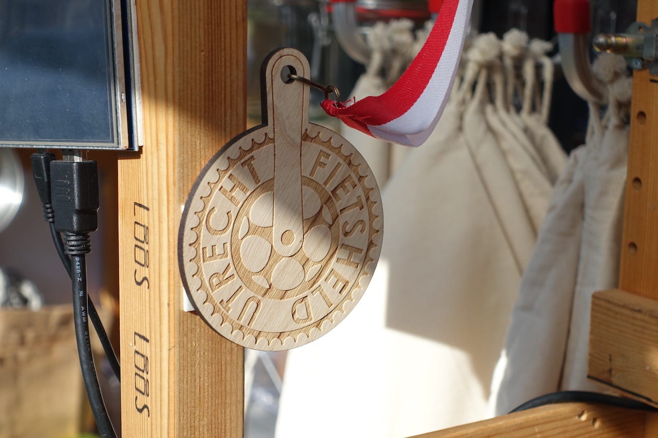 A small wooden medal, with a red and white ribbon attached. The medal is affixed to the inside of the LOOS trailer. On the medal is a motif of a gear and crank arm from a bicycle. Around the outside it reads Fietsheld Utrecht