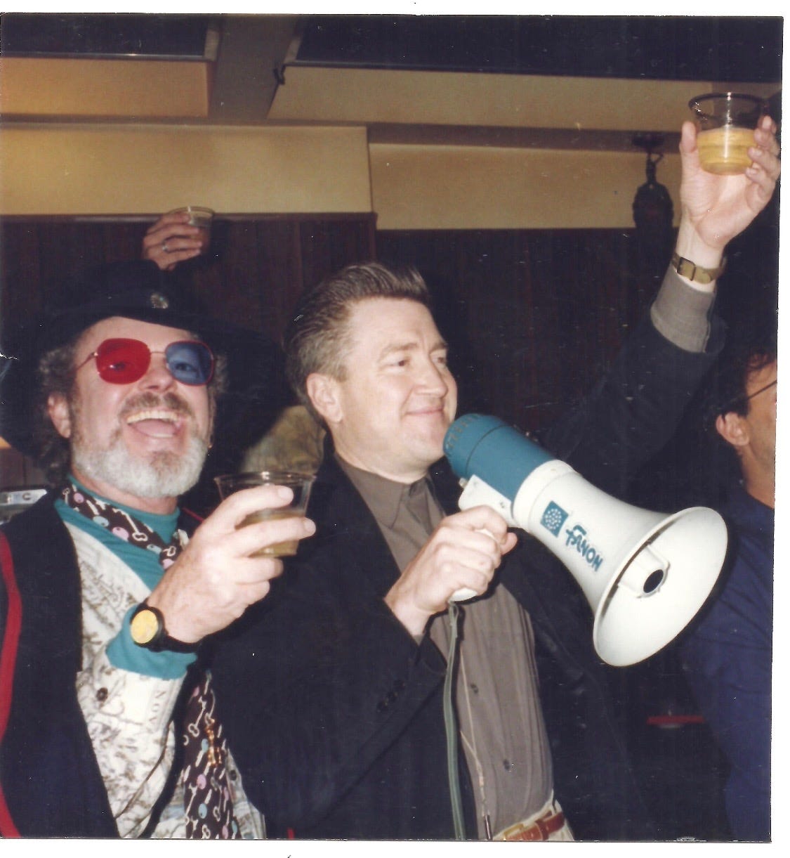 A digital photo taken of a film photo. A young David Lynch and Russ Tamblyn are at a party. Russ stands next to David as David gives a toast into a megaphone. Russ looks at the camera. He wears Dr. Jacoby's signature glasses. David, Russ, and other partygoers all have their plastic cups raised in celebratory "cheers" to David's toast.