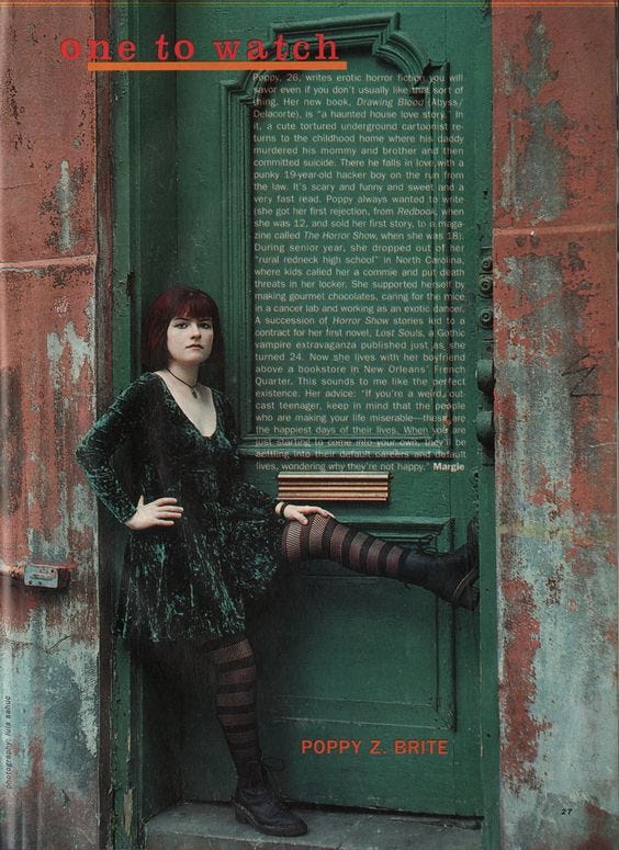 A magazine page captioned One to Watch. Text is illegible. Photo shows Poppy Z. Brite, an auburn haired woman in a dark green crushed velvet baby doll dress leaning up against a rusty looking old wall