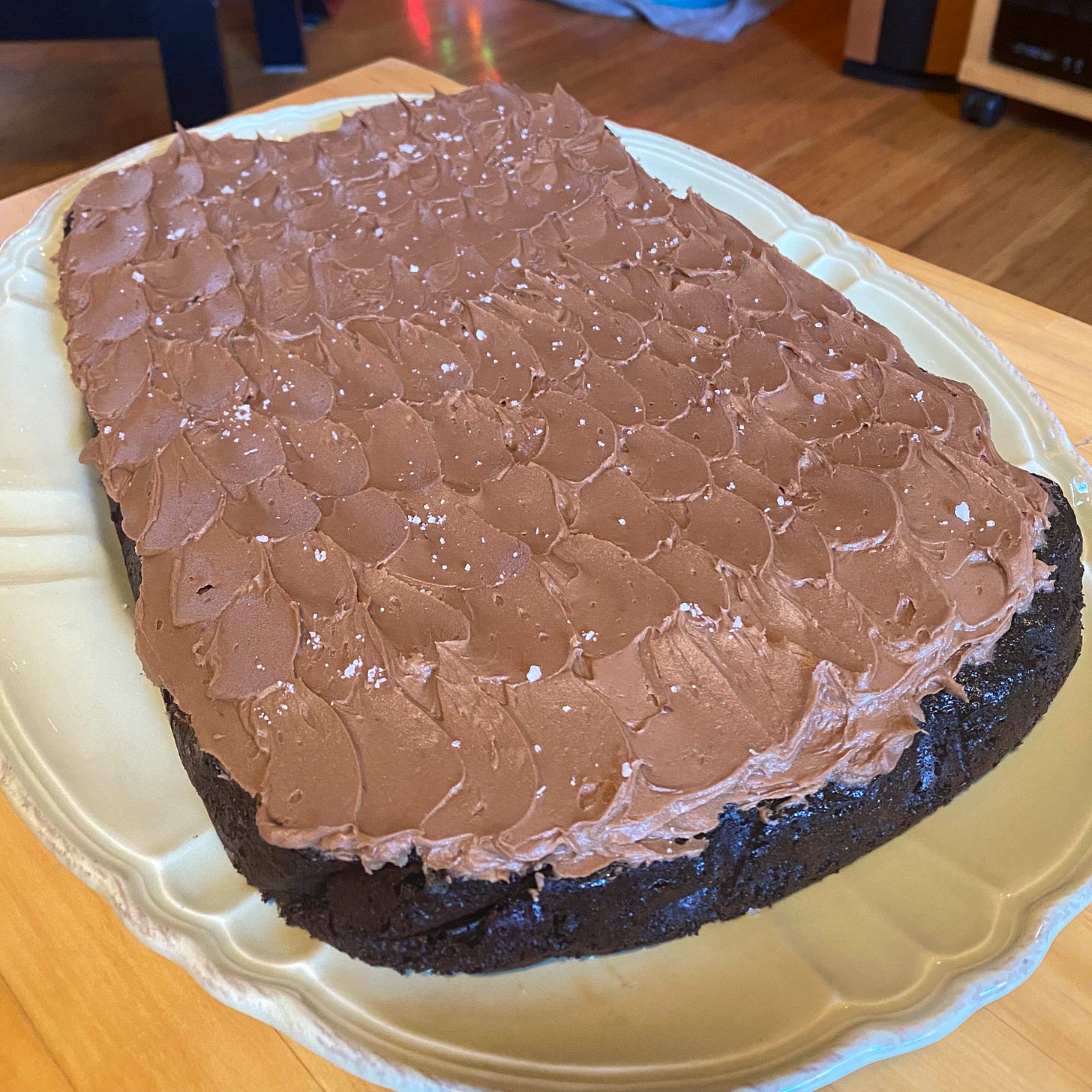 A large chocolate sheet cake with milk chocolate icing in waves, sprinkled with coarse salt and set on a green serving plate on the coffee table.