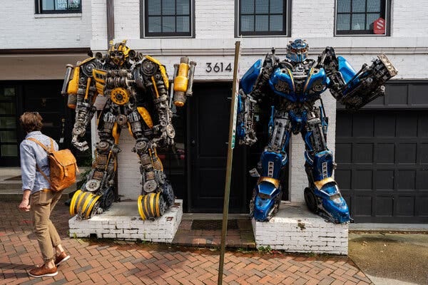 Two large “Transformers” sculptures stand on platforms outside a white-brick townhouse.
