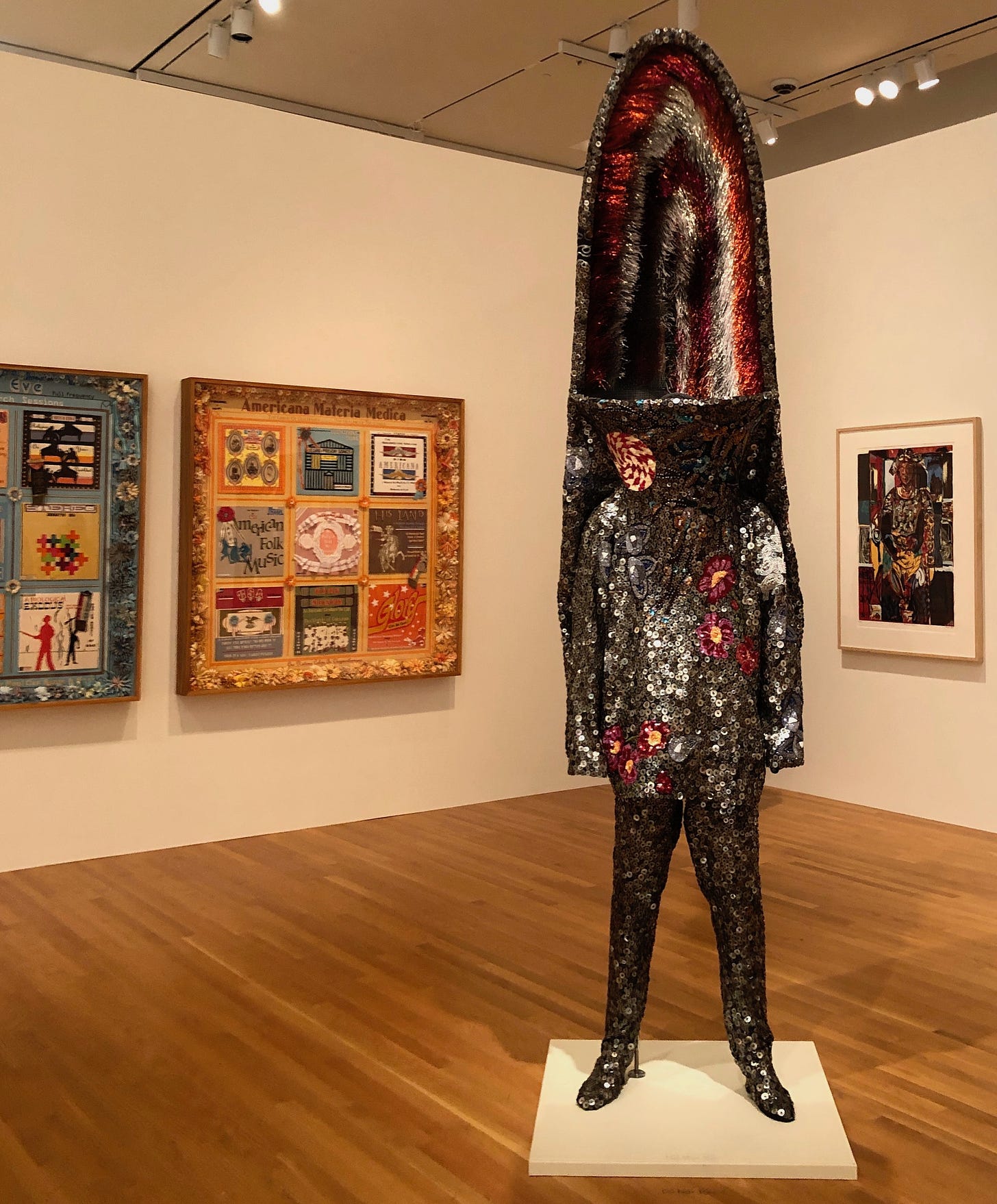 A glistening, full-body costume with an enormous headdress, in a museum gallery. Three artworks are on the walls behind.