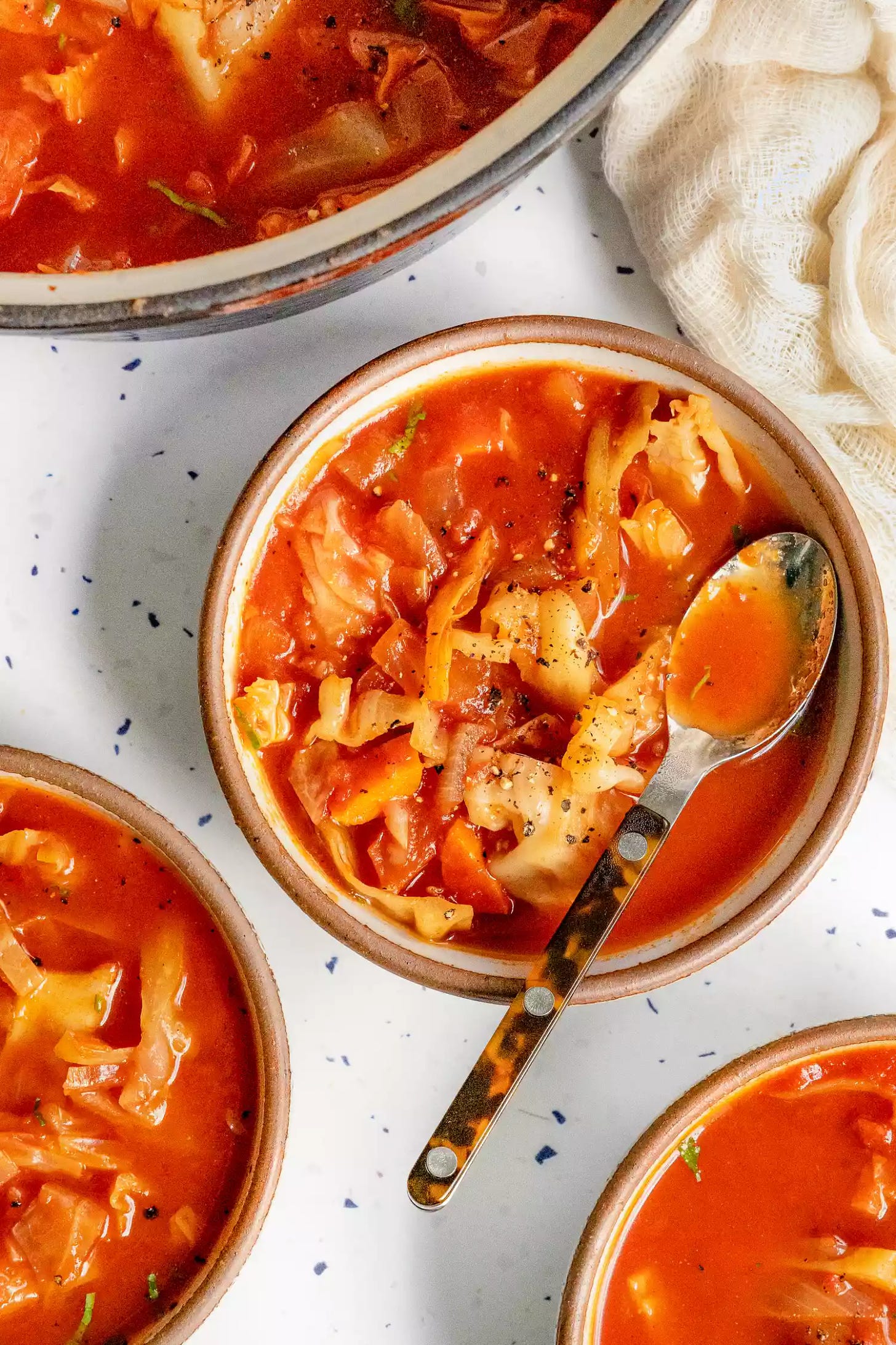 Sweet and Sour Cabbage Soup in Three Bowls with Spoons, and Next to It, a Large Pot with More Soup and a Kitchen Linen