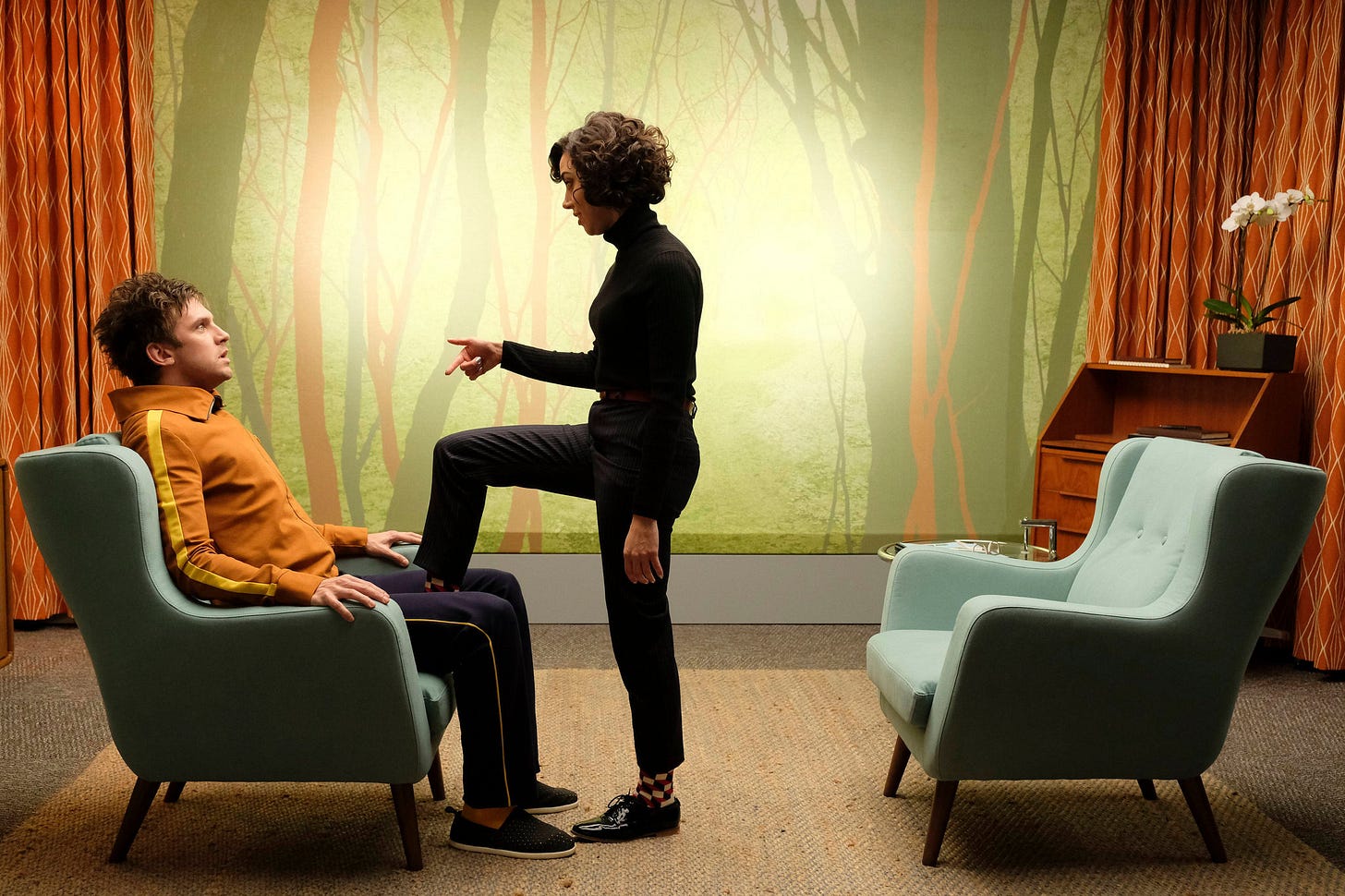 a white guy in an orange sweatshirt sits in a chair; a woman stands in fron tof him with her foot between his legs lecturing him