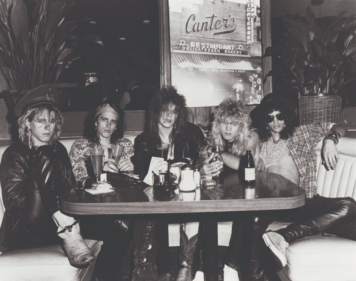 Image: Guns N’ Roses at Canter’s Deli, Los Angeles, CA, c.1980s. Photo by Jack Lue
Image description: A black and white photo of the ive young men from Guns n' Roses rock n' roll band are seated at a table under a sign that reads "Canter's." They are styled with long hair in the 1980s fashion of "hair metal" bands. On the far right, Slash wears his signature hat and sunglasses.
