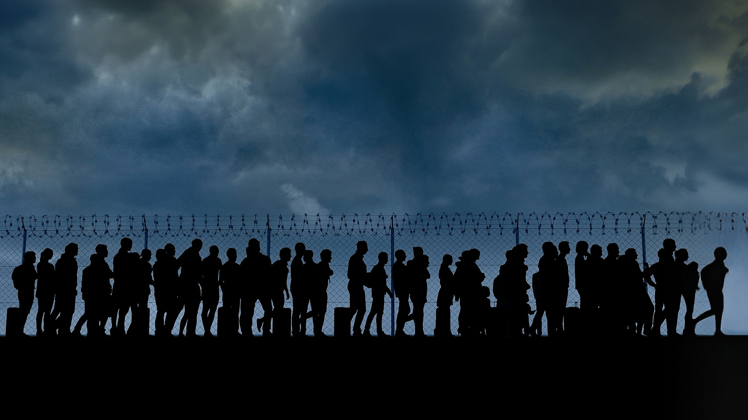 Dark silhouettes of people along a barbed wire fence with a cloudy sky