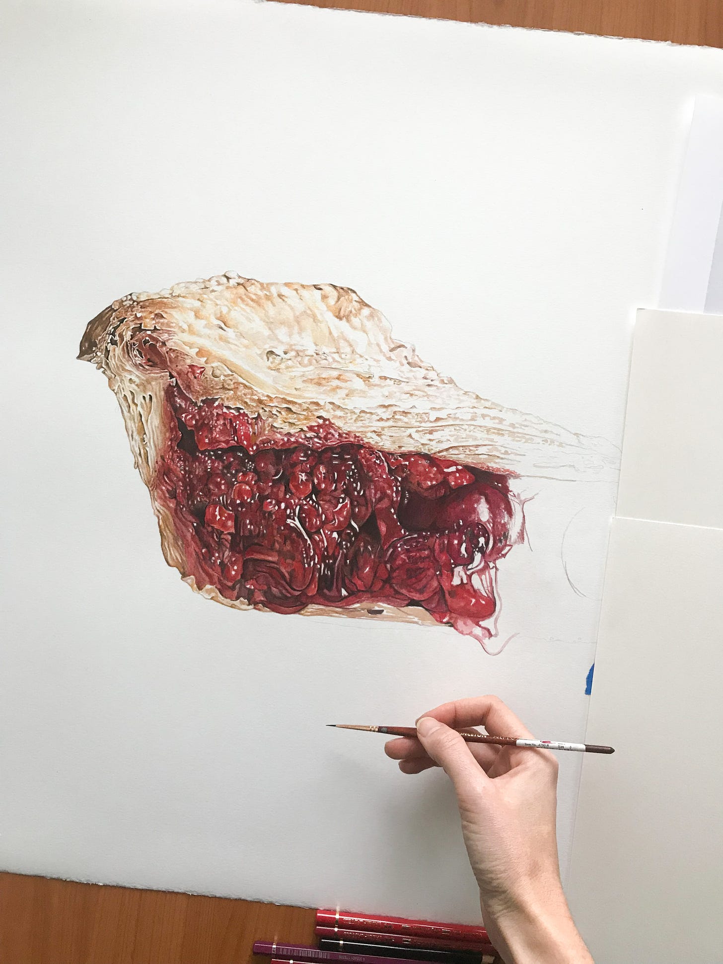 A birdseye view of the cherry pie painting, mid painting, with my hand in the bottom corner with a paintbrush. You can see the pencil lines below the pie, and where the paint is layered