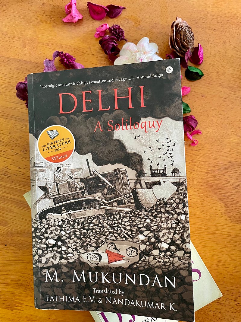 Image of the book, Delhi: A Soliloquy by M Mukundan lying on the table with dried flowers and another book, City of Djinns by William Dalrymple.