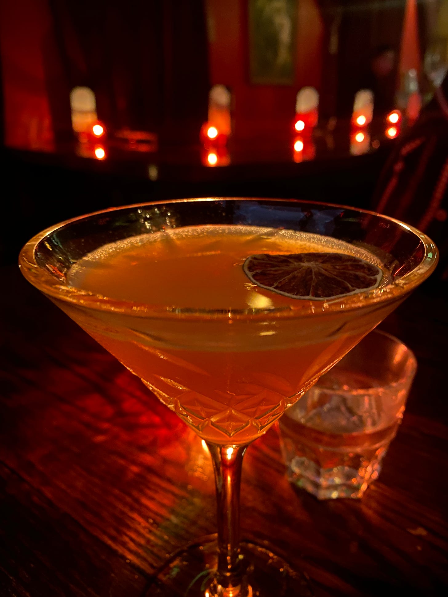 a martini glass with embossed diamond shapes near the stem, filled with an orange-brown drink that has a dried lime slice floating on top. The room in the background is dimly lit by candles, making everything seem red-hued.