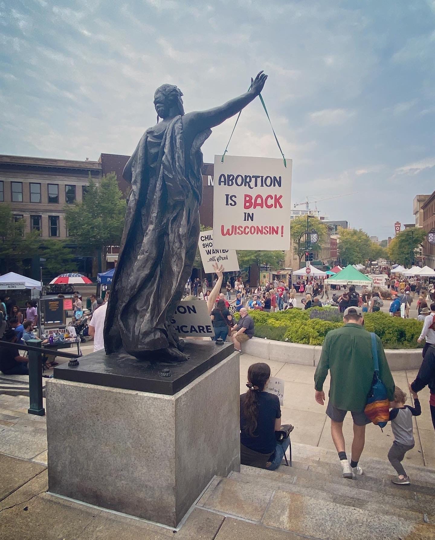 A hand-painted sign hangs from the outstretched hand of a bronze statue of a woman overlooking a crowded State Street in Madison. The sign reads, "Abortion is back in Wisconsin!"