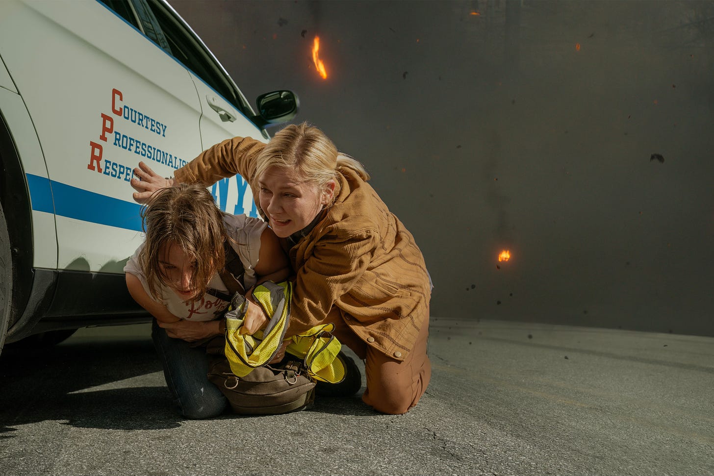 Kirsten Dunst shields Cailee Spaeny from the aftermath of an explosion against the side of a police car. Flames are still licking the air, and both women look shaken.