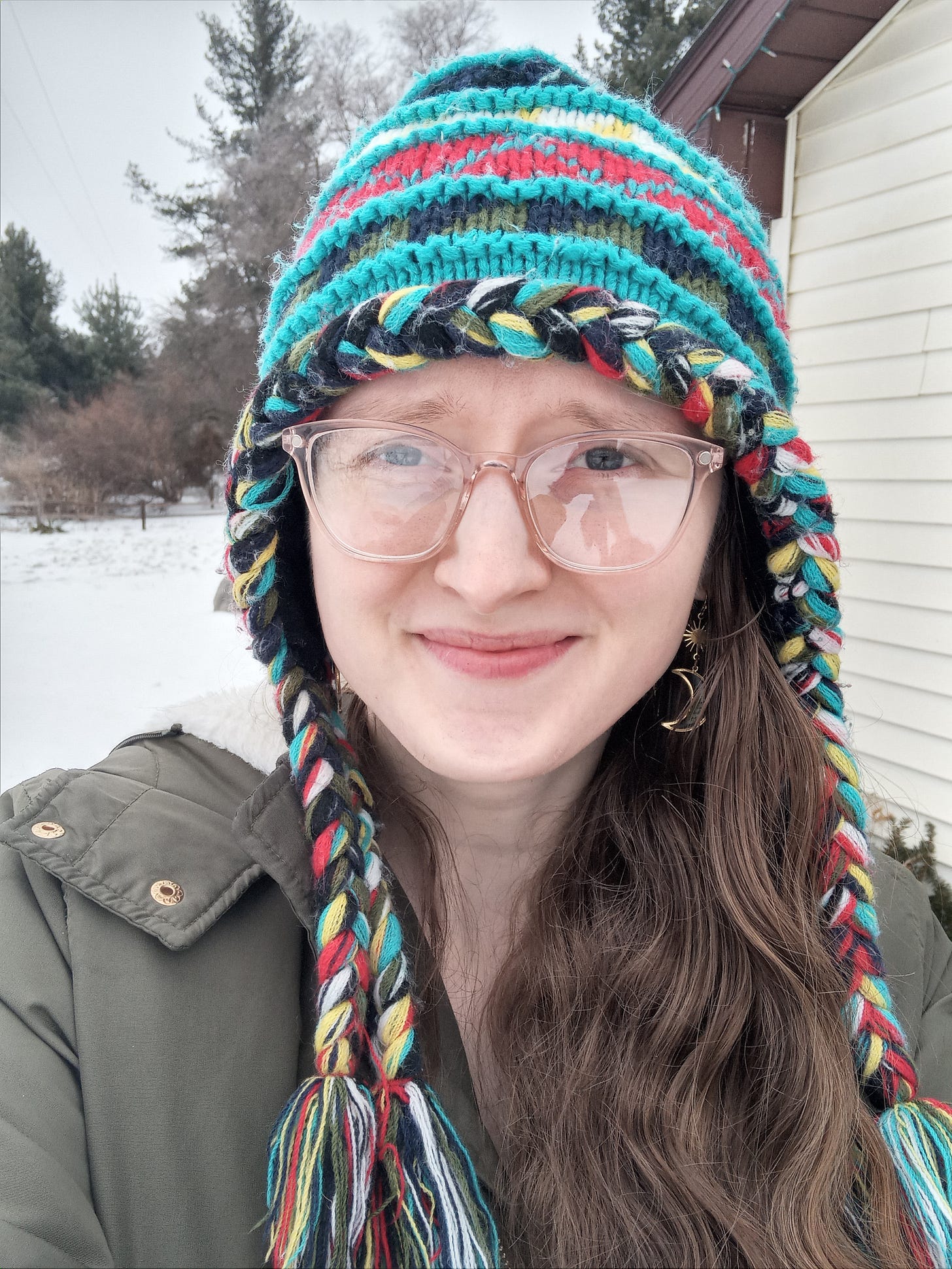 Selfie of Kandi Zeller (She/Her), a white woman with long reddish brown hair and pink glasses, wearing a green winter coat and a colorful winter hat made of acrylic yarn, against a backdrop of snow and evergreen trees