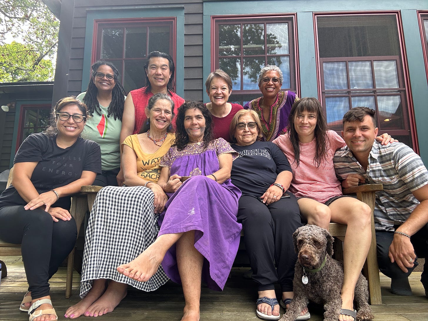 Group photo on a porch of 12 smiling people in casual clothes. Black, white, asian, latino and one grey poodle. All middle aged or older. Big smiles.