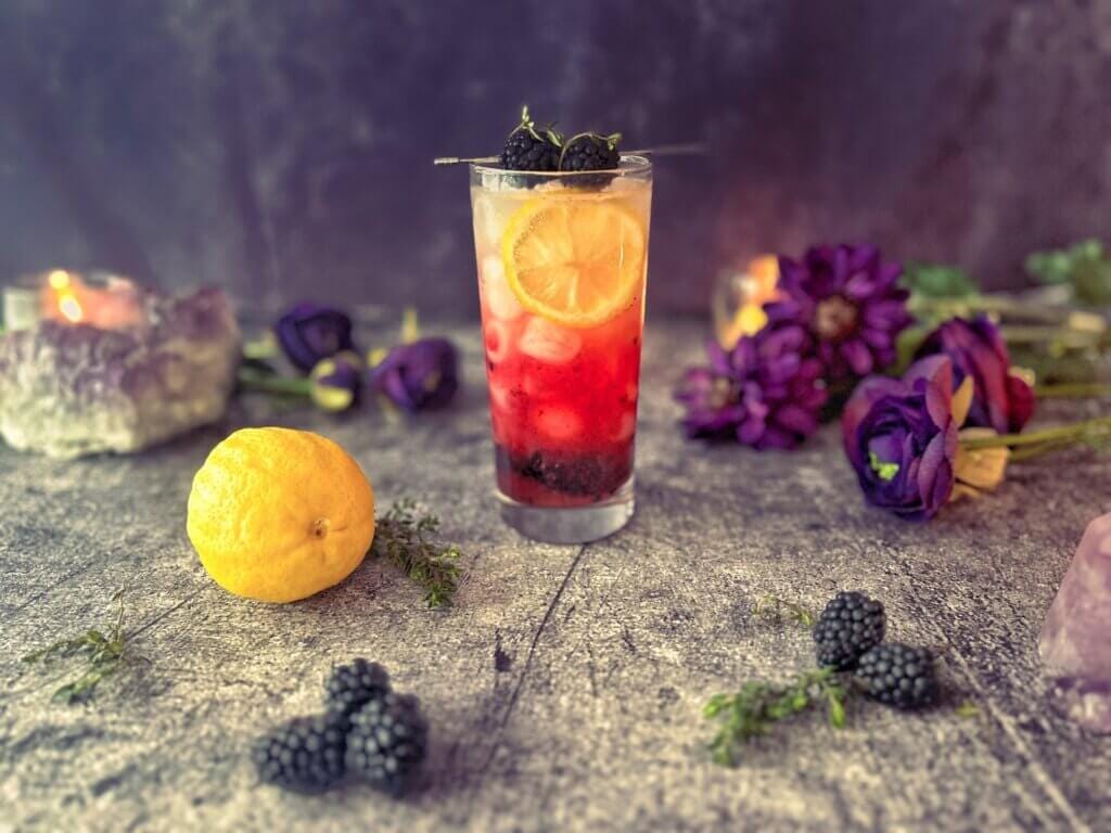 A purple and gold mocktail in a tall skinny glass, garnished with a lemon wheel and skewered blakcberries