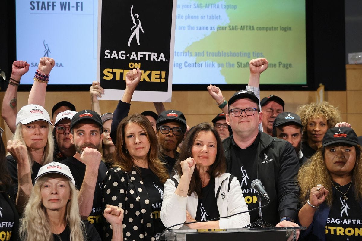 A still from the SAG-AFTRA press conference. Duncan Crabtree-Ireland stands behind a podium. Next to him is SAG-AFTRA President Fran Drescher. Around them stand many union members wearing SAG-AFTRA shirts holding up their fists. Michelle Hurd stands in the right hand corner of the image. 